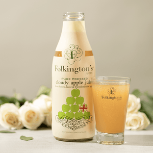 Open bottle of 1 litre Apple Juice alongside a Folkington's glass of apple juice on a counter. There is a bunch of white roses and green leaves laying in the background behind the bottle and glass.