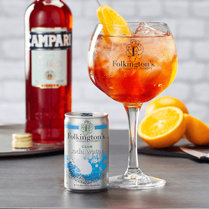 Lifestyle image of a can of Folkington's 150ml Soda Water with a bottle of Campari open in the background. Next to the can is a glass of Campari and Soda Water with ice and a slice of Orange to garnish.